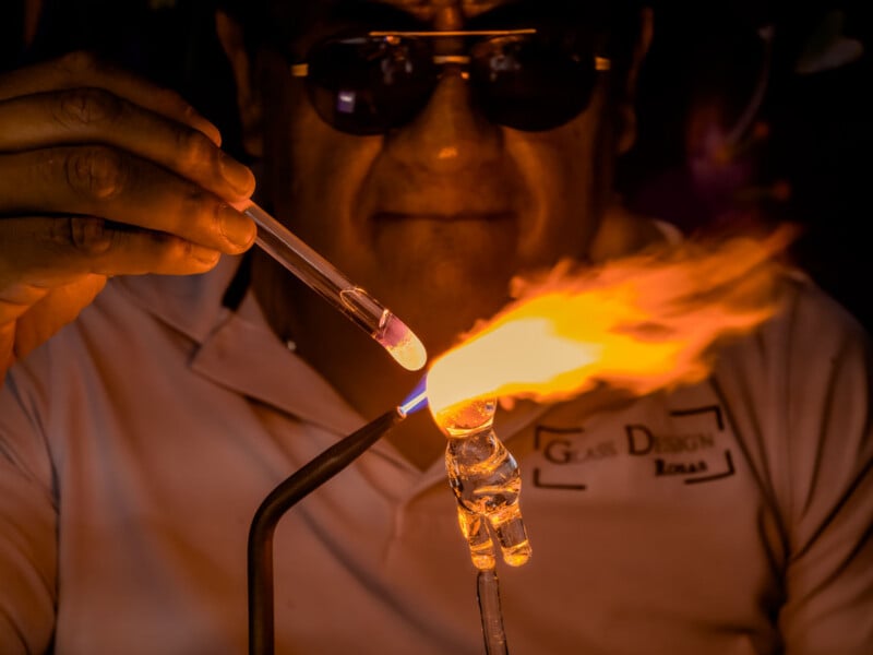 A person wearing sunglasses uses a torch to heat glass, shaping a small figurine while holding a glass rod and tube. Bright flames illuminate their hands and the partially finished glass sculpture.