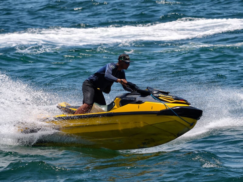A person wearing a cap and sunglasses rides a yellow jet ski at high speed on choppy blue water, creating white spray and wake behind them.