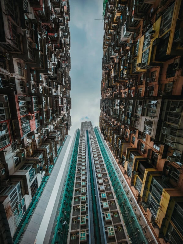 A dramatic upward view between two tall, colorful, densely packed apartment buildings, framing a modern high-rise skyscraper against a cloudy sky.