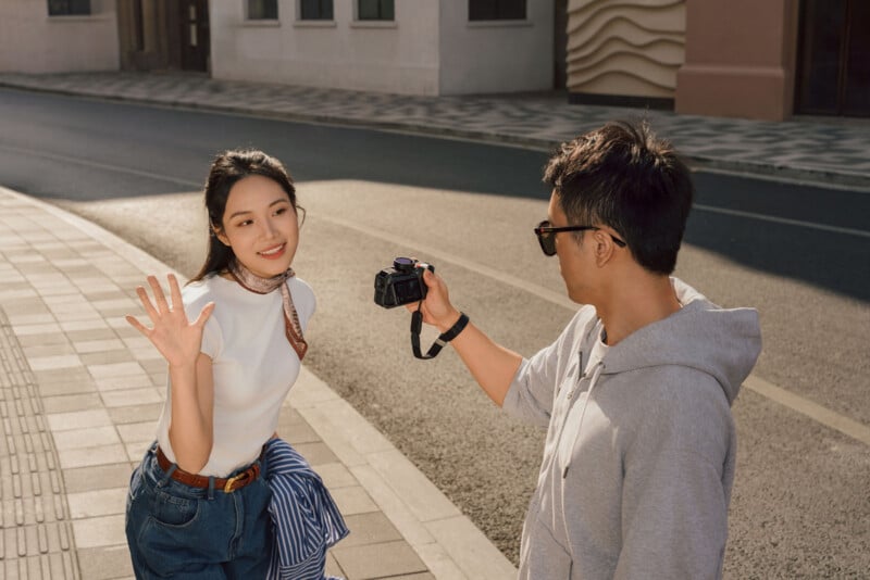 A woman smiles and waves at the camera while a man in sunglasses photographs her on a sunny street. The woman stands near the sidewalk, and the street and buildings are visible in the background.