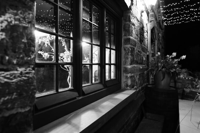 Black-and-white photo of a stone building at night, featuring a window reflecting indoor flowers, string lights above, and a flower pot on a barrel outside. The scene has a cozy, rustic atmosphere.