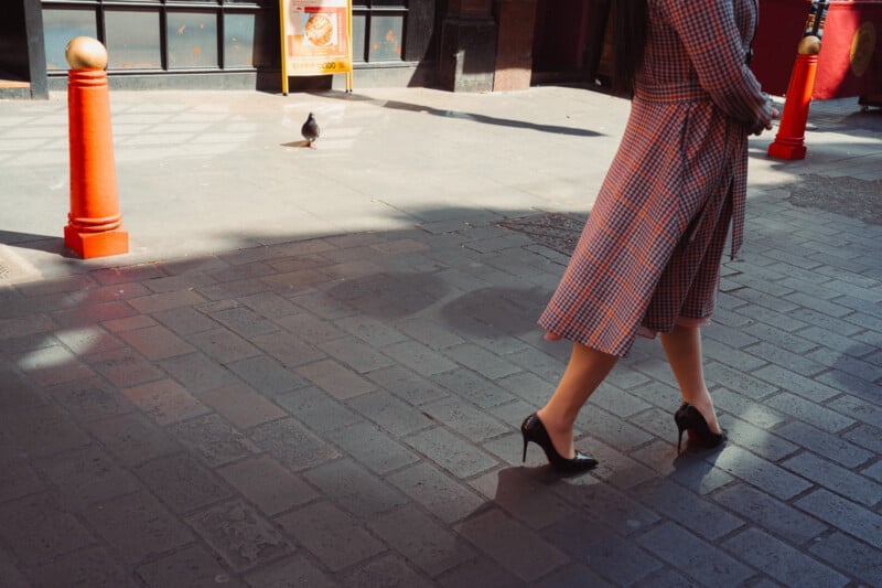 A person in a checkered dress and black high heels walks on a sunlit cobblestone street. Red bollards and a pigeon are visible in the background. Only the lower half of the person is shown.