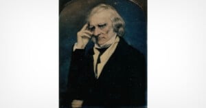 An older man with white hair sits in a dark suit, raising his right hand to his temple in a thoughtful pose, set against a dark background.