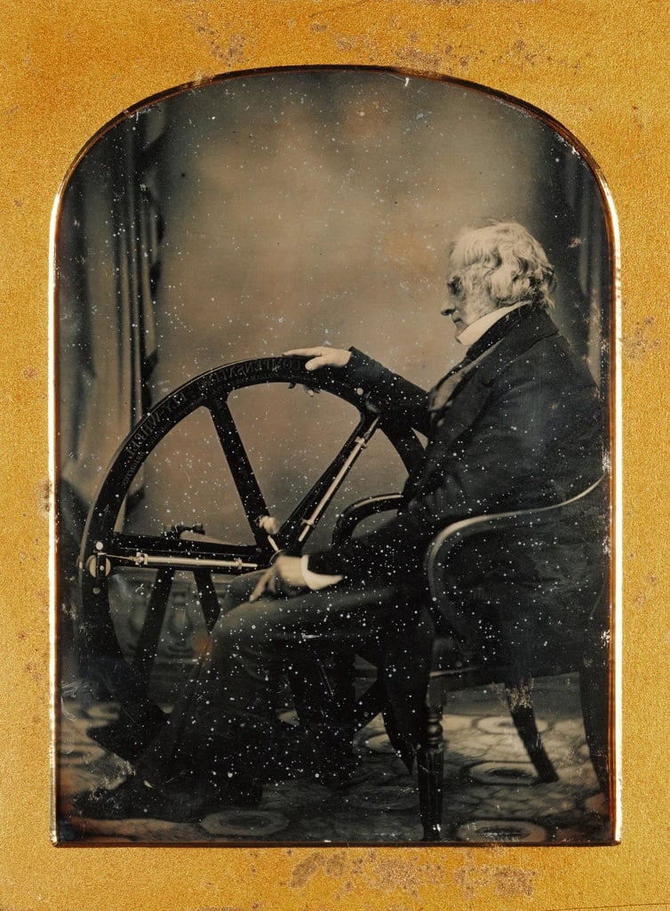 A sepia-toned vintage photograph of an elderly man sitting in a chair, facing sideways, with his hand resting on a large mechanical wheel. The image is framed with a gold border.