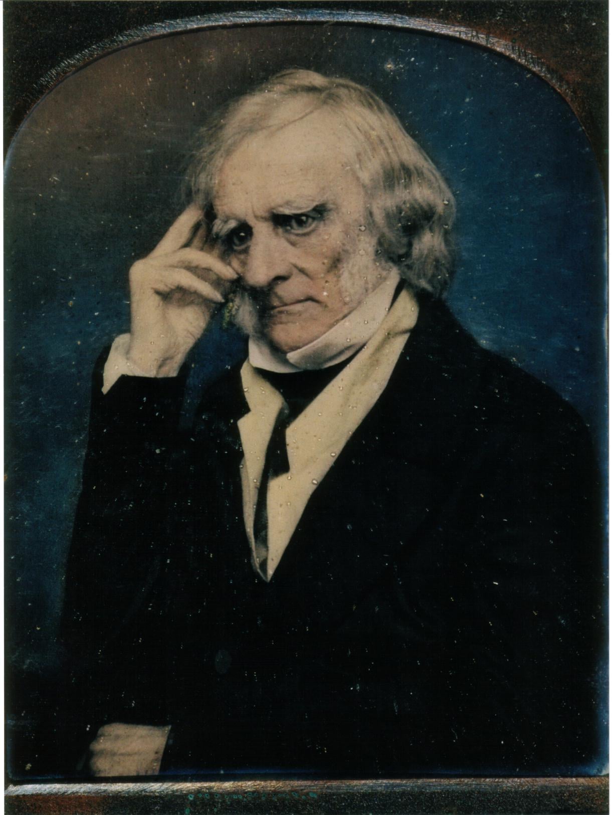 An elderly man with white hair and sideburns, wearing a dark suit and cravat, sits with one hand resting on his temple and a serious expression, posing against a blue and brown background.