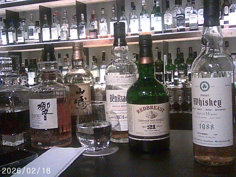 A selection of whiskey and whisky bottles, including Hibiki, Yamazaki, BenRiach, Redbreast, and an Irish whiskey, displayed on a bar counter with a glass of water and shelves of bottles in the background.