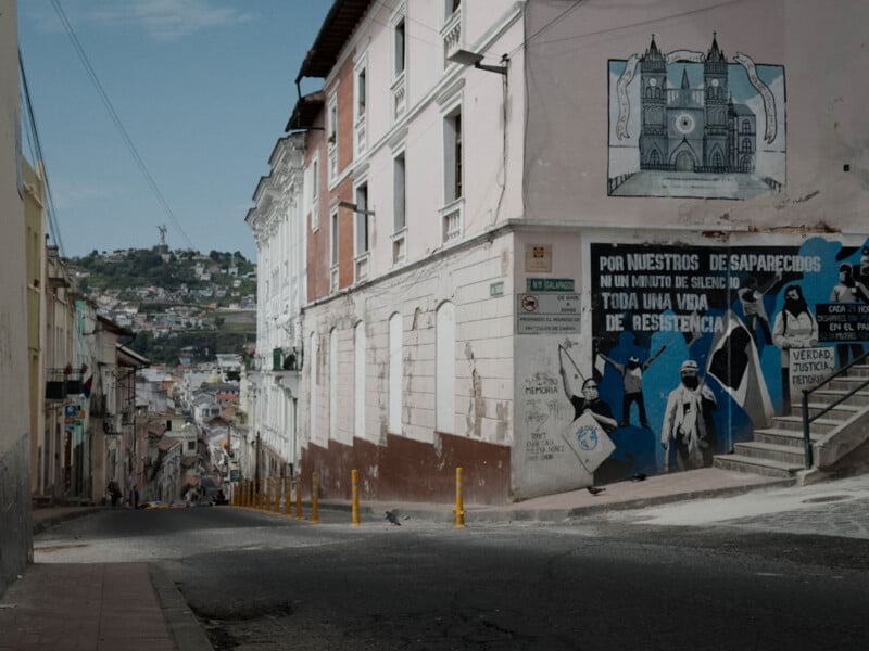 A quiet, steep street lined with old buildings, colorful murals, and political graffiti. In the background, a distant statue stands atop a green hill under a partly cloudy sky.