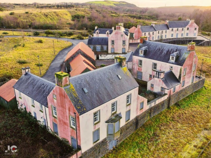 A cluster of empty, weathered pink buildings, likely abandoned, stands surrounded by overgrown grass and hills under a cloudy sky, connected by roads and stone walls.