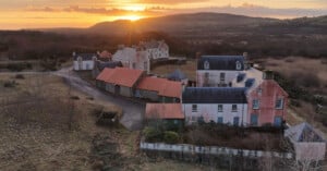 A cluster of rustic buildings with red roofs and weathered walls sits in a rural landscape at sunset, surrounded by fields and hills under a warm, glowing sky.