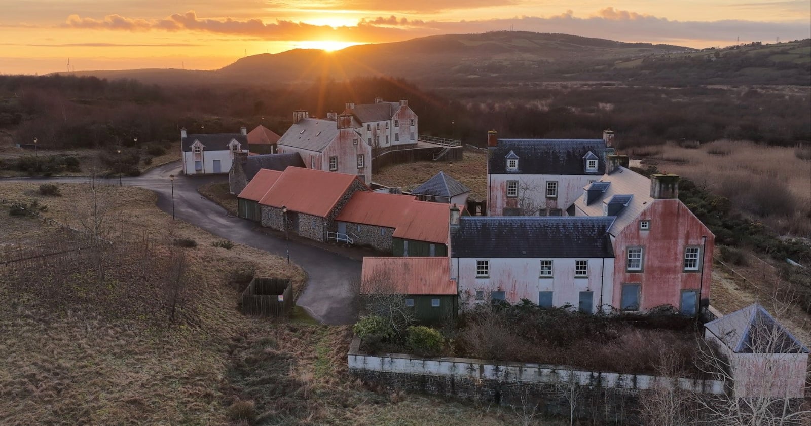 A cluster of rustic buildings with red roofs and weathered walls sits in a rural landscape at sunset, surrounded by fields and hills under a warm, glowing sky.