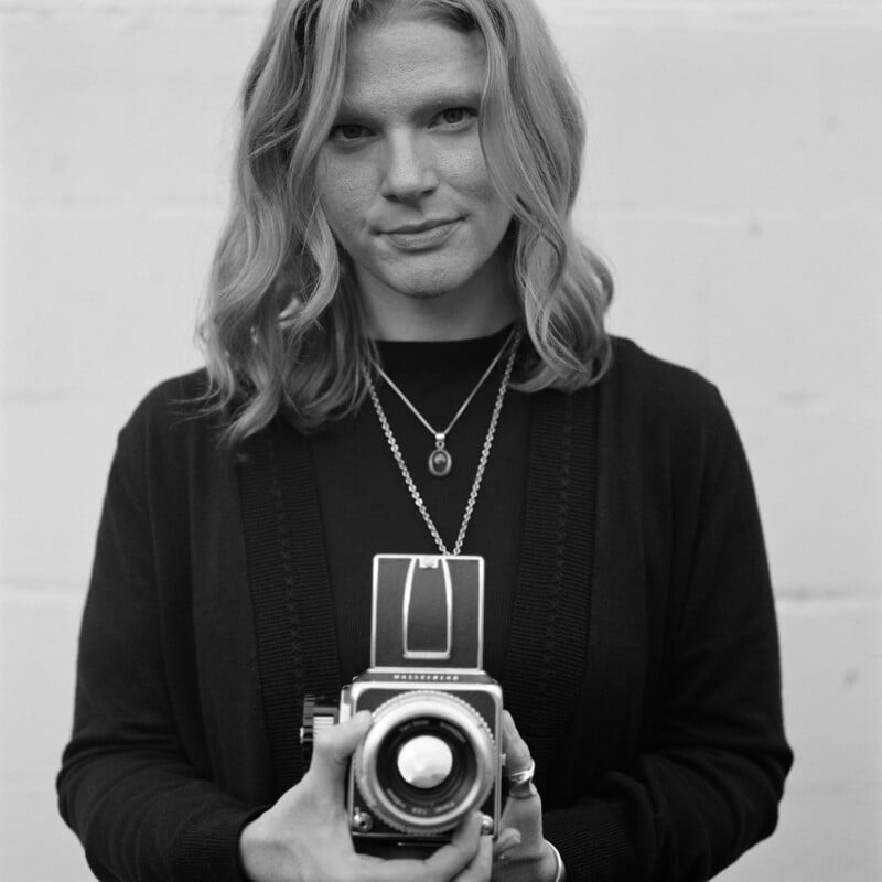 A woman with shoulder-length wavy hair holds a vintage camera and looks at the viewer. She wears a dark sweater and layered necklaces, standing against a plain, light-colored wall. The image is in black and white.