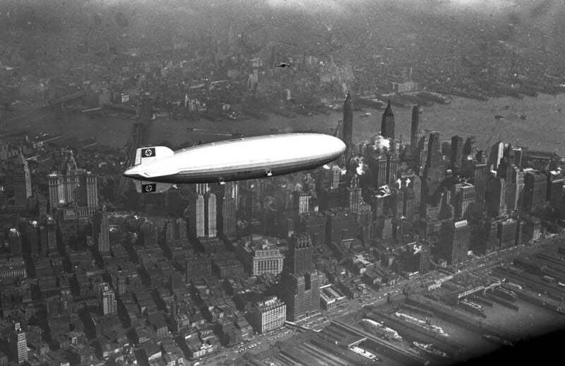 A large airship with swastika emblems on its tail flies over Manhattan, New York City, with skyscrapers and the Hudson River visible below on a hazy day.