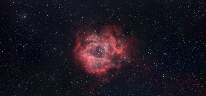A deep space image of the Rosette Nebula, featuring a large, reddish-pink cloud of interstellar gas and dust surrounded by a field of stars against the blackness of space.