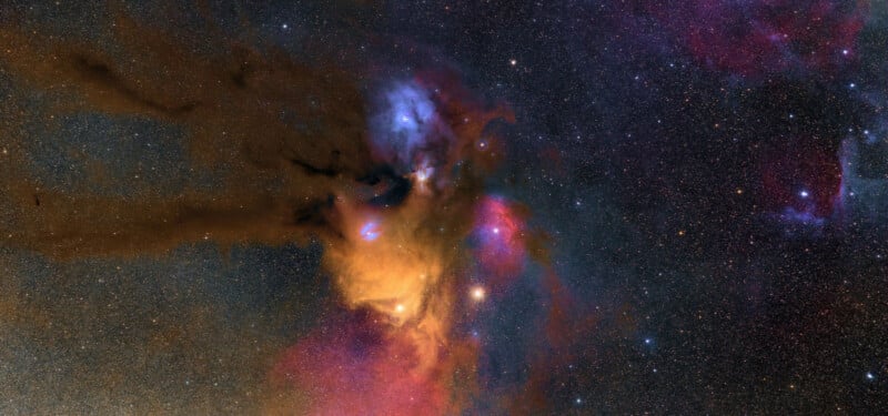 A vibrant deep space scene showing a colorful nebula with swirling clouds of orange, blue, and purple dust, surrounded by countless bright stars against a dark sky.