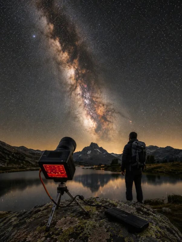A person stands by a lake under a star-filled night sky with the Milky Way visible. A camera on a tripod faces the sky, capturing the scene. Mountains and reflections are visible in the background.