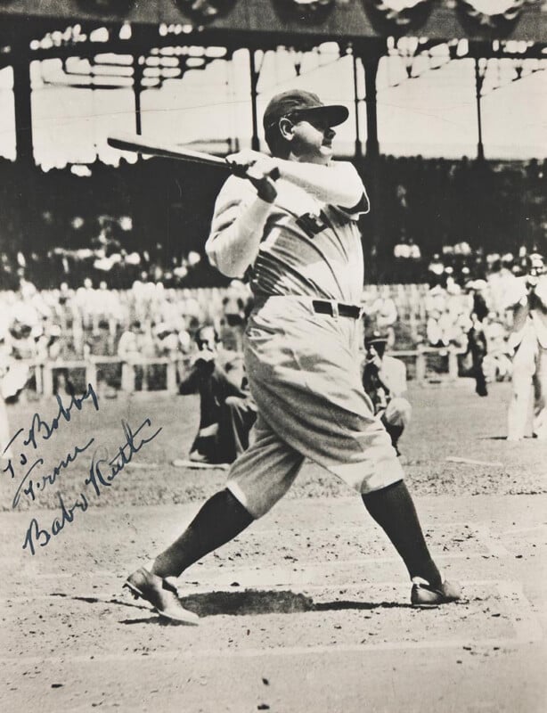 A vintage black-and-white photo of a baseball player mid-swing at home plate, wearing a classic uniform and cap, with spectators and a stadium visible in the background. An autograph is written on the bottom left.