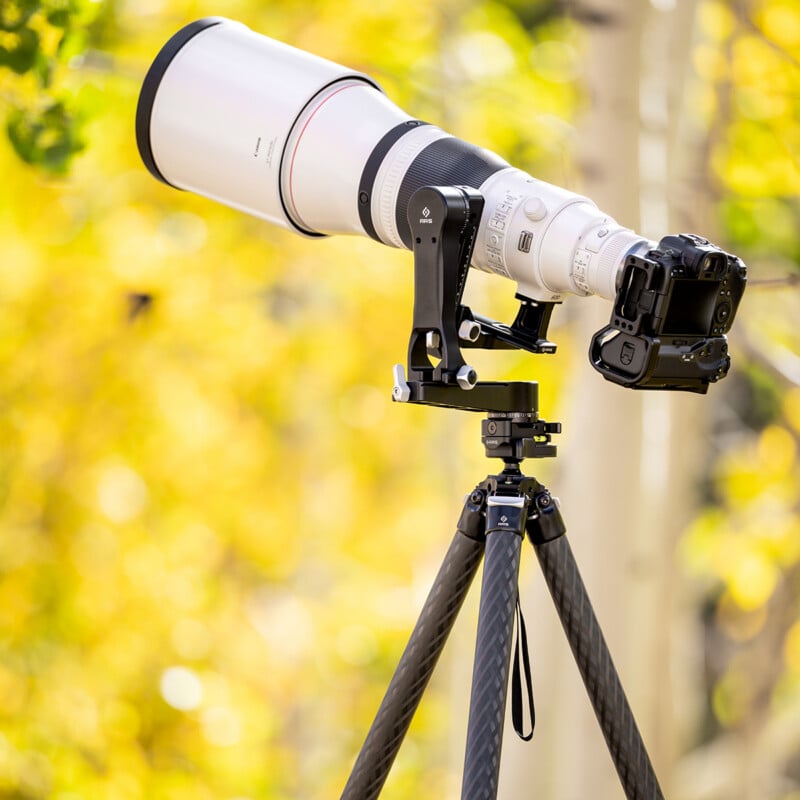 A professional camera with a large telephoto lens mounted on a sturdy tripod, set outdoors with a blurred background of yellow and green foliage.