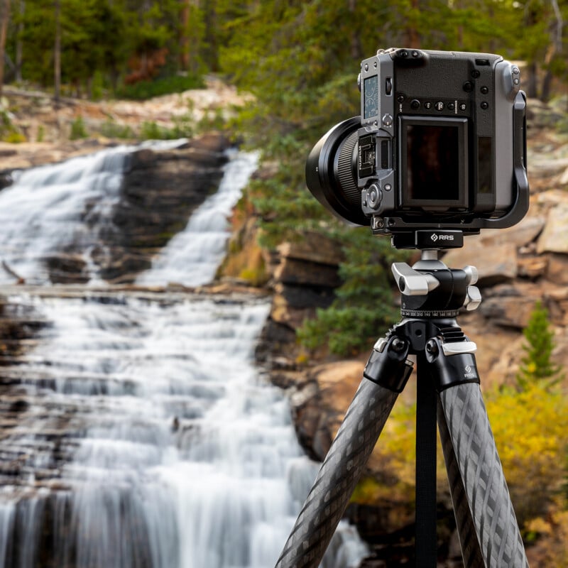 A camera on a tripod is set up outdoors, facing a blurred cascading waterfall surrounded by rocks and green trees, capturing the scenic landscape.