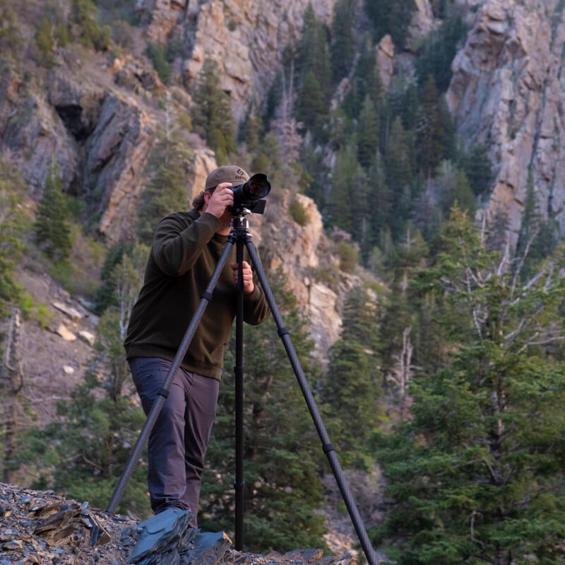 A person in outdoor clothing uses a camera on a tall tripod, standing on rocky ground with a forest and steep, rocky mountains in the background.
