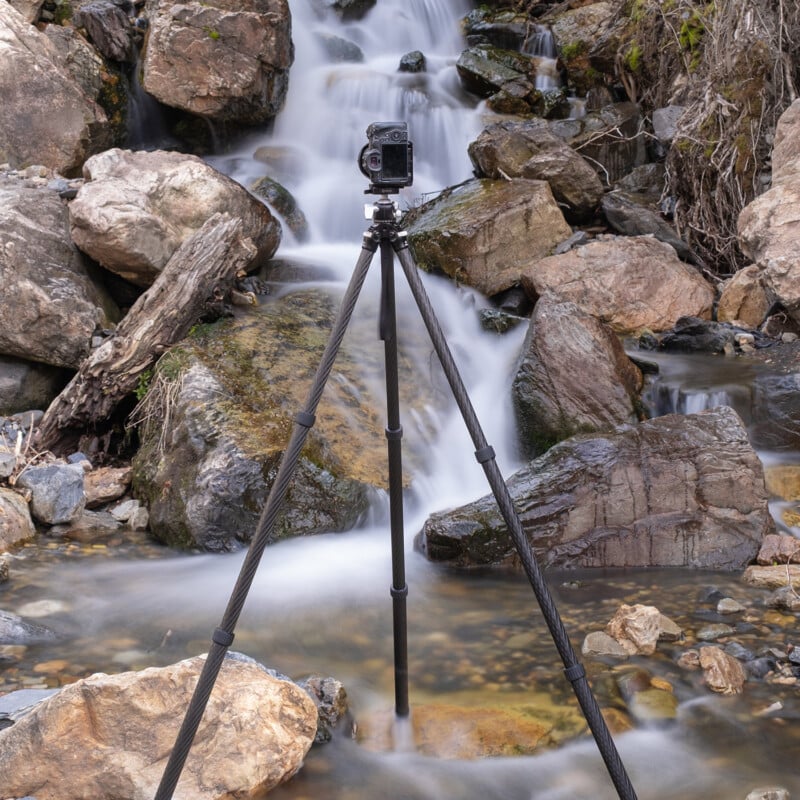 A camera mounted on a tripod is set up in front of a small waterfall, surrounded by rocks and flowing water in a natural outdoor setting.