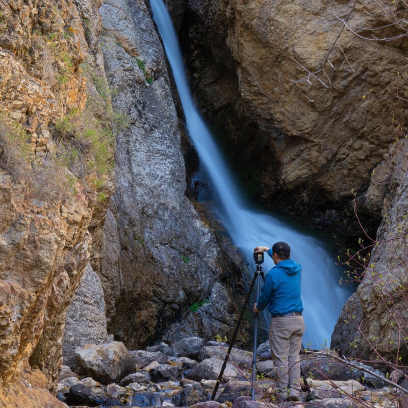 A person in a blue jacket uses a camera on a tripod to photograph a waterfall cascading down rocky cliffs in a natural, outdoor setting.