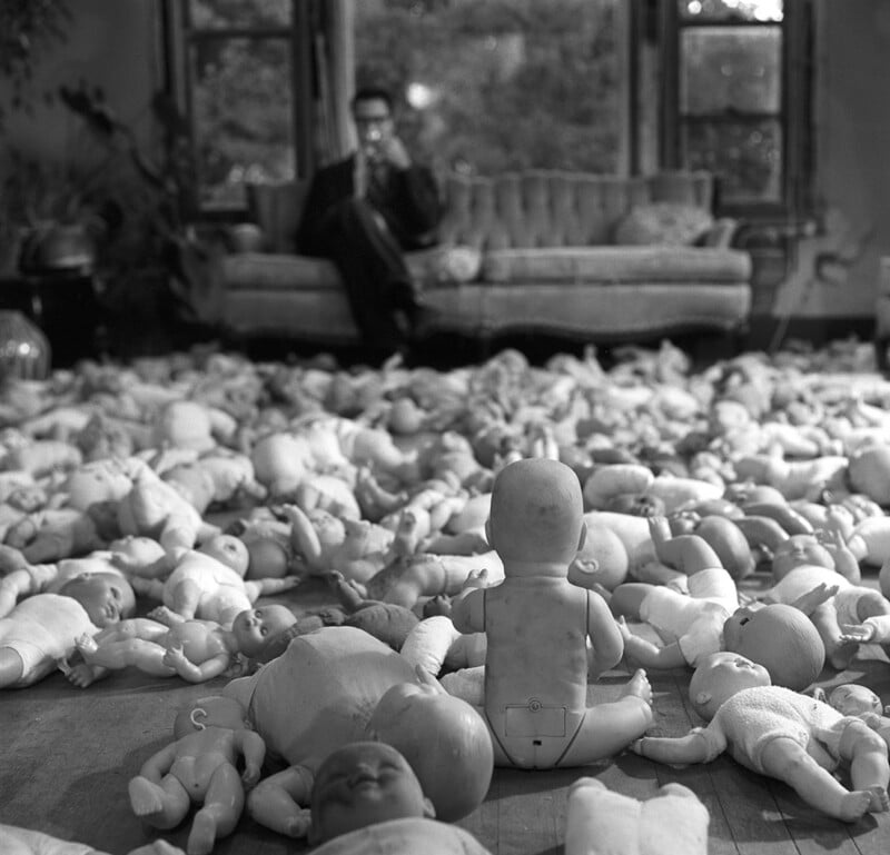 A black and white photo shows a living room floor covered with numerous disassembled dolls. In the background, a person in a suit sits on a couch, partially blurred, while the dolls are clearly in focus in the foreground.