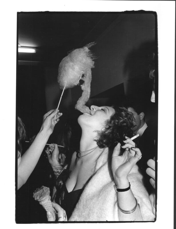 A woman in evening attire leans back with her mouth open, about to eat cotton candy held by someone else. She holds a cigarette in one hand and an ice cream cone in the other, surrounded by people at a lively gathering.