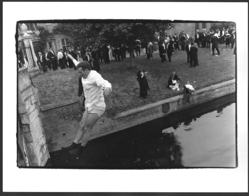 A man in a shirt and underwear jumps into a river from a stone edge, while people on the grassy bank behind him watch, gather, and take photos. Some are dressed formally, suggesting a celebratory event.