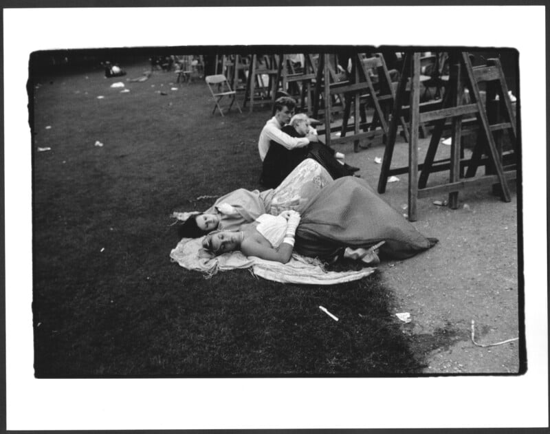Four people in formal clothing rest on blankets on the grass, with several wooden chairs in the background. The scene suggests the end of an outdoor event, with scattered litter on the ground.