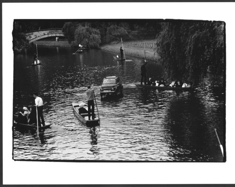Black and white photo of people punting on a river; a car is partially submerged in the water, surrounded by several punts, with a stone bridge and trees in the background.