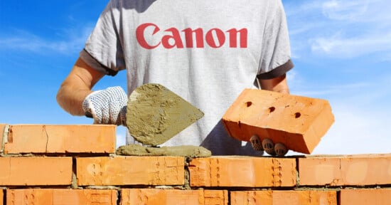 A person in a gray Canon T-shirt builds a brick wall, holding a trowel with mortar in one hand and a brick in the other, under a bright blue sky.