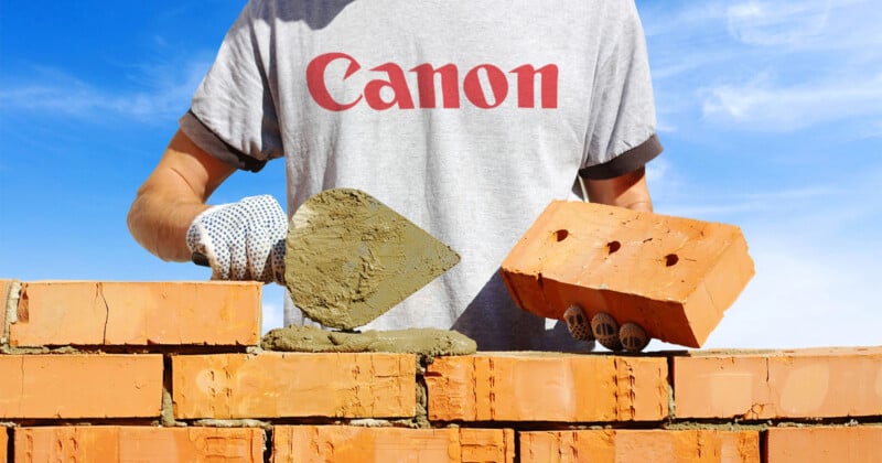 A person in a gray Canon T-shirt builds a brick wall, holding a trowel with mortar in one hand and a brick in the other, under a bright blue sky.