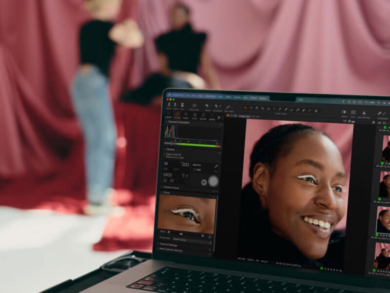 A photo editing software is open on a laptop, displaying close-up portraits of a smiling person with white eyeliner. In the blurred background, two people appear to be setting up a photoshoot in a studio with pink drapes.