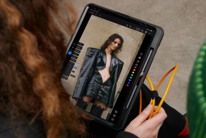 A person holds a tablet displaying a photo of a woman wearing a black leather jacket and skirt, standing against a beige wall. The tablet shows editing tools and colorful markers on the screen.