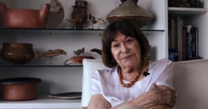 An older woman with short brown hair sits on a white chair, wearing a white shirt and a necklace. Behind her are shelves displaying various ceramic pots and animal figurines. She is looking at the camera with a gentle smile.
