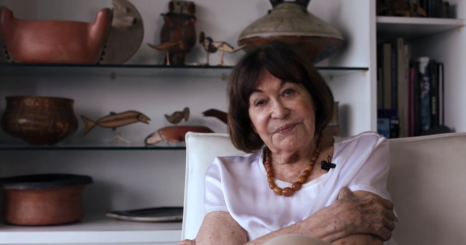 An older woman with short brown hair sits on a white chair, wearing a white shirt and a necklace. Behind her are shelves displaying various ceramic pots and animal figurines. She is looking at the camera with a gentle smile.