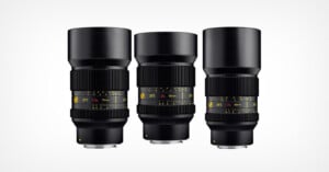Three black camera lenses with yellow and white markings are standing upright side by side against a plain white background. Each lens displays different focal lengths and aperture information on their barrels.