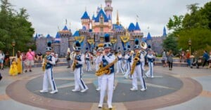 A marching band in white and blue uniforms performs with brass instruments in front of Disneyland’s Sleeping Beauty Castle, surrounded by visitors and colorful decorations.