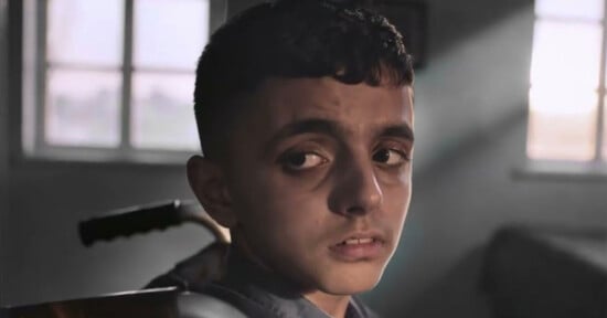 A boy with short dark hair looks over his shoulder with a serious expression. Soft light from a window behind him partially illuminates his face in a dimly lit room.