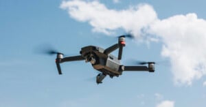A black quadcopter drone with a camera hovers in the sky, its propellers spinning. The background features a blue sky with a few white clouds.