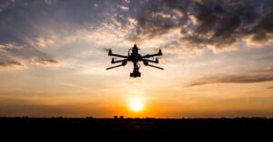 A large drone with multiple rotors flies in silhouette against an orange and yellow sunset sky, with scattered clouds and the sun low on the horizon.