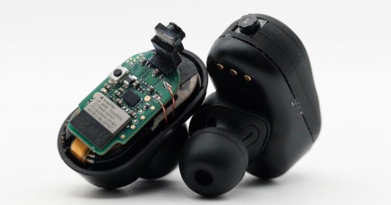 Two black wireless earbuds, one of which is opened to reveal its internal green circuit board and electronic components, while the other earbud remains intact with visible charging contacts.