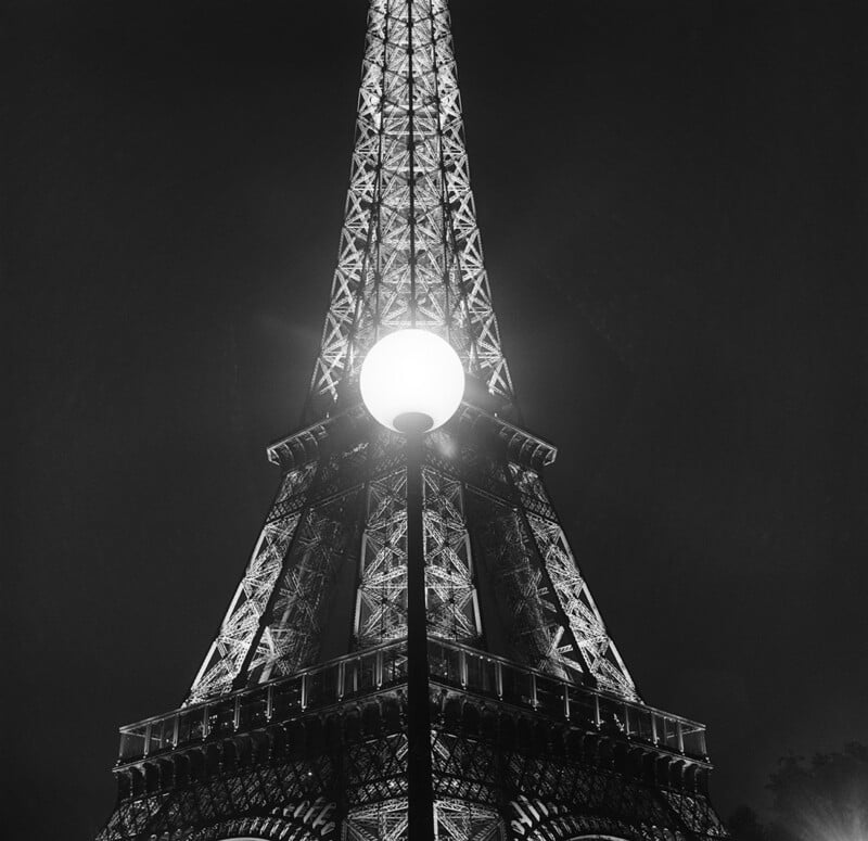 Black and white photo of the Eiffel Tower at night, viewed from below with a glowing streetlamp centered in the foreground, partially obscuring the tower's illuminated structure.