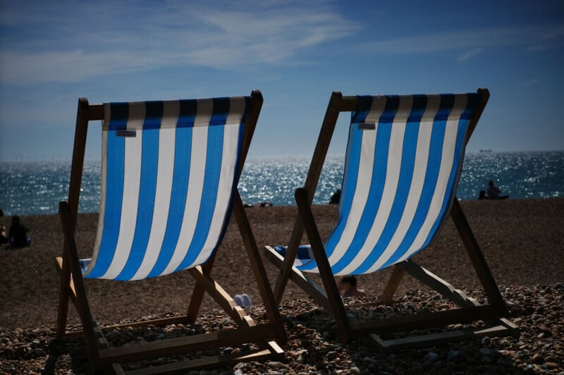 Two empty blue-and-white striped deck chairs sit on a pebbly beach, facing a sparkling sea under a clear blue sky. Silhouettes of people and a distant ship can be seen in the background.