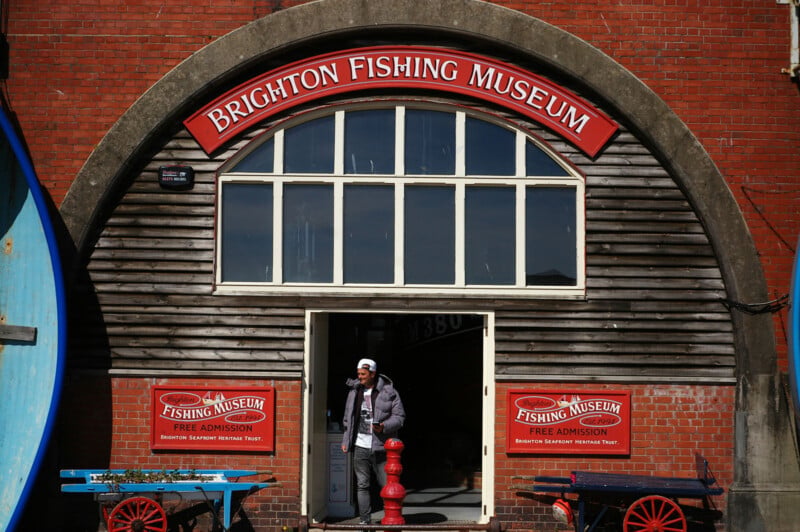 A person stands in the doorway of the Brighton Fishing Museum, a brick building with a large arched sign above the entrance and red benches with flower boxes on either side of the door.