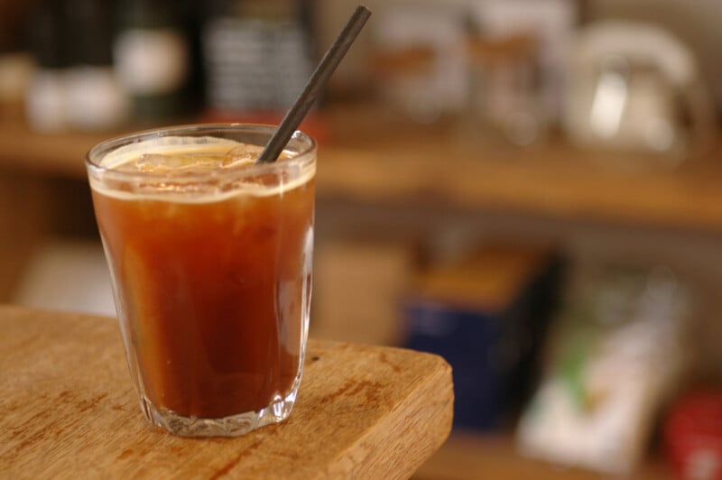 A glass of iced coffee with a black straw sits on a wooden surface, with a blurred background of shelves and various objects.
