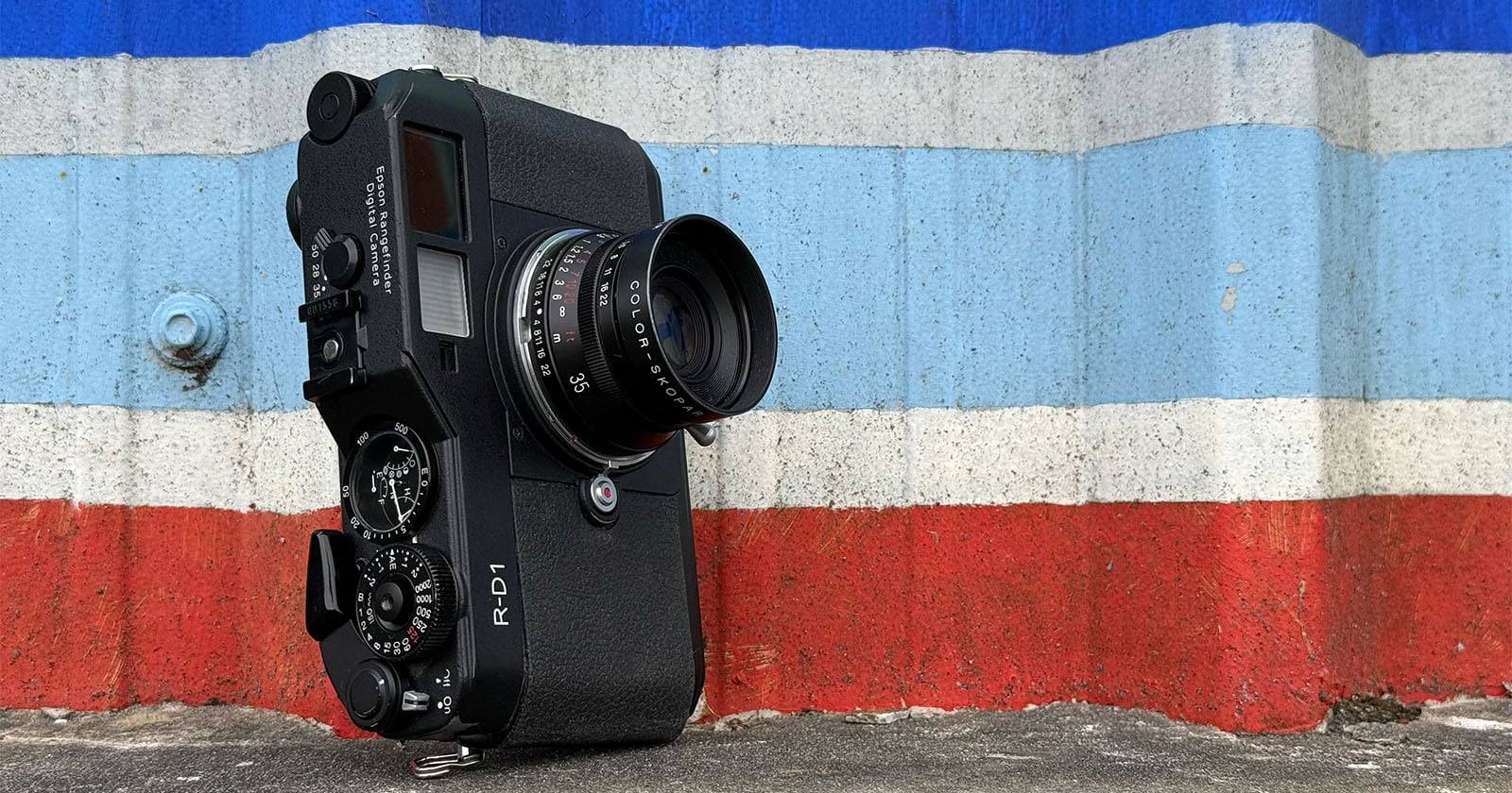 A black RD-1 rangefinder camera with a lens is resting upright on a concrete surface in front of a wall painted with horizontal blue, white, and red stripes.