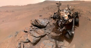 A Mars rover sits on rocky, reddish terrain surrounded by scattered boulders and dusty hills under a hazy sky, capturing the barren Martian landscape.