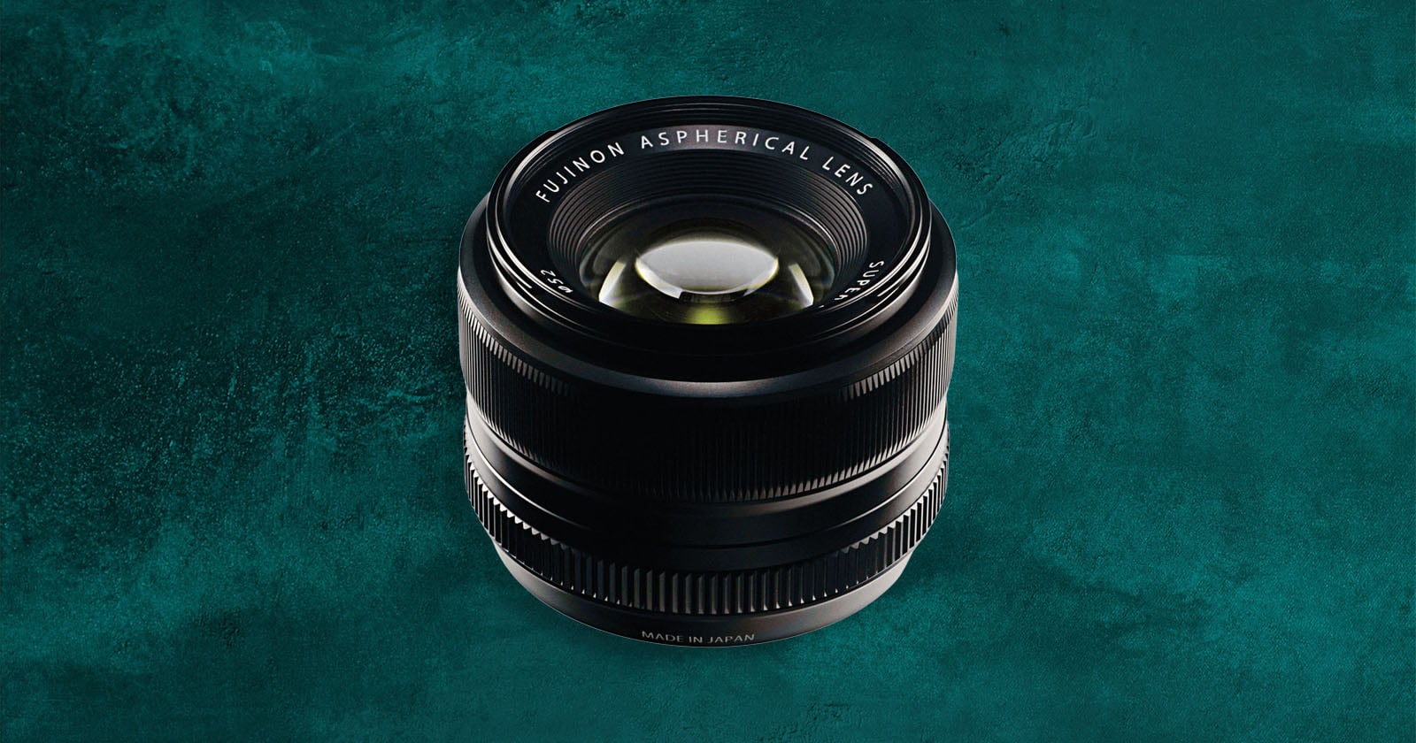 A Fujinon aspherical camera lens is positioned upright on a textured teal background, with "Fujinon Aspherical Lens" and "Made in Japan" visible on the lens barrel.