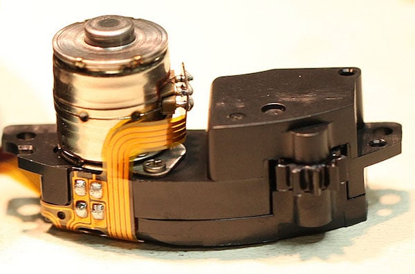 Close-up of a small electric motor with a metallic cylindrical component attached to a black plastic housing, connected by yellow flexible circuit wiring.